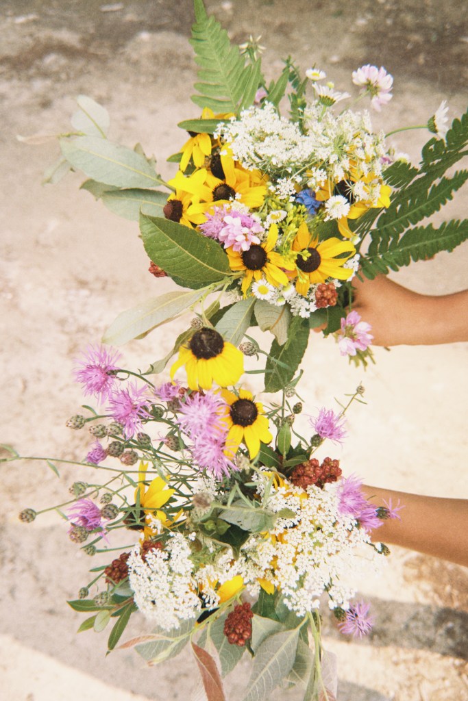 girls at a lake in the upper peninsula of Michigan holding a a bouquet of flowers at one of their favorite camping spots to stay at before a big bus event called Skoolie U.P. In the upper peninsula of Michigan. By Adventurous Leila.