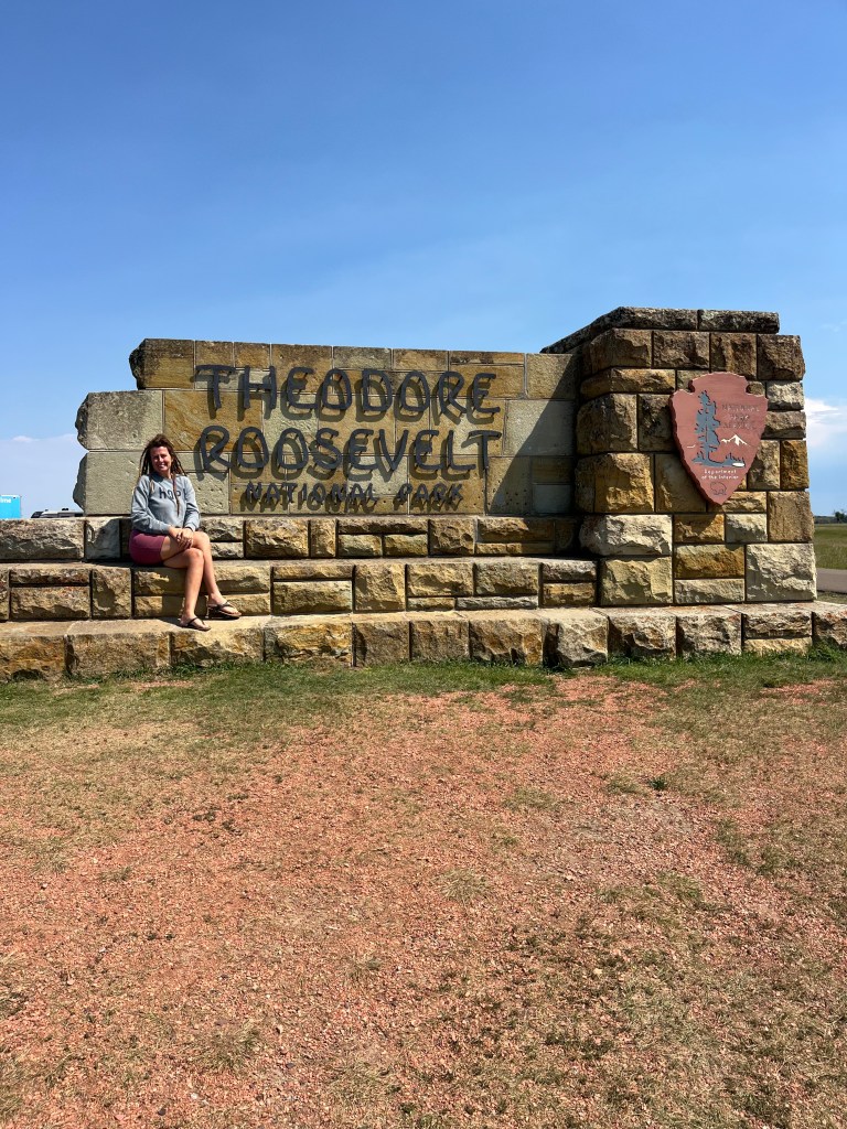 A girl sitting in front of theater Roosevelt national Park sign. By Adventurous Leila.